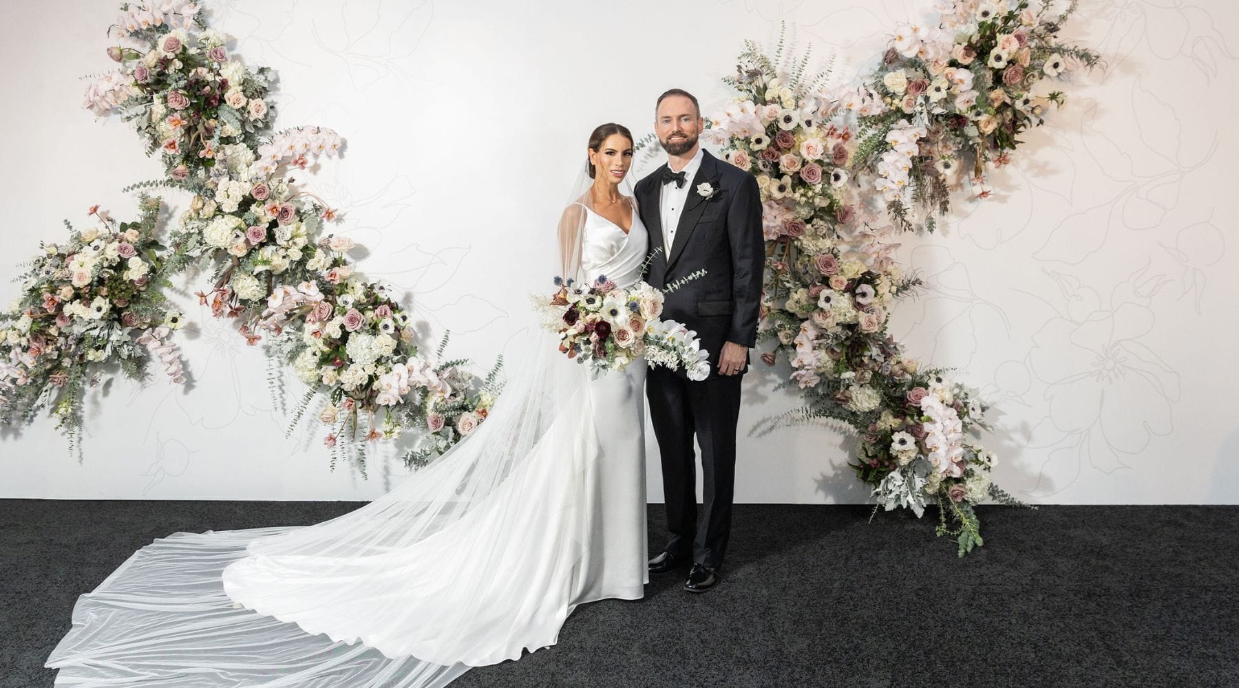 Newlywed Couple Posing in Front of An Elegant Floral Backdrop. The Bride Wears a Sleek White Gown With a Long Train and Holds a Bouquet of Flowers, While the Groom Stands Beside Her in A Classic Black Tux. - dbandrea